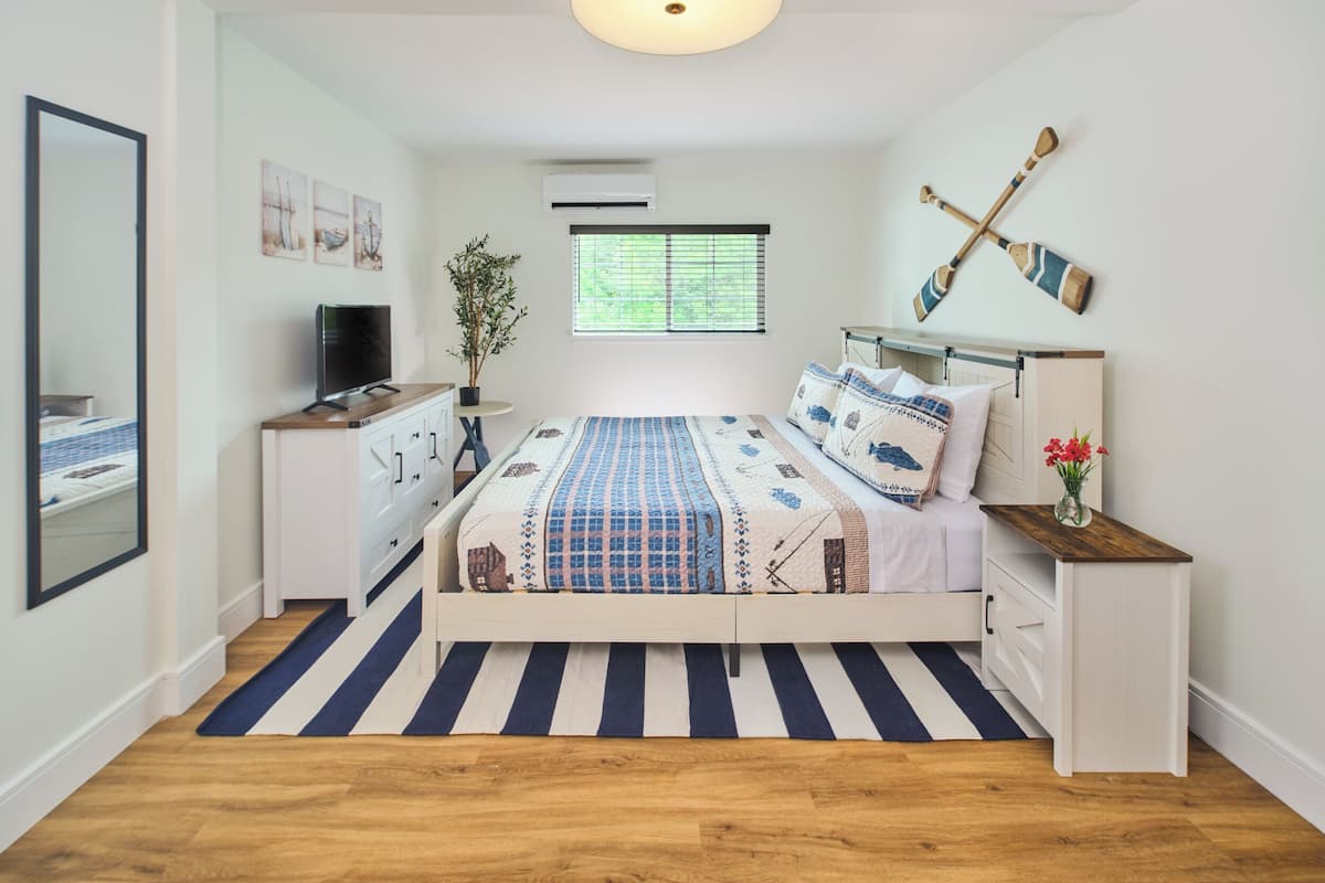 Un dormitorio con cama, mesitas de noche, televisor, planta y ventana con persianas.