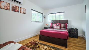 A bedroom with a large bed, two windows with blinds, a wooden floor, and a rug.