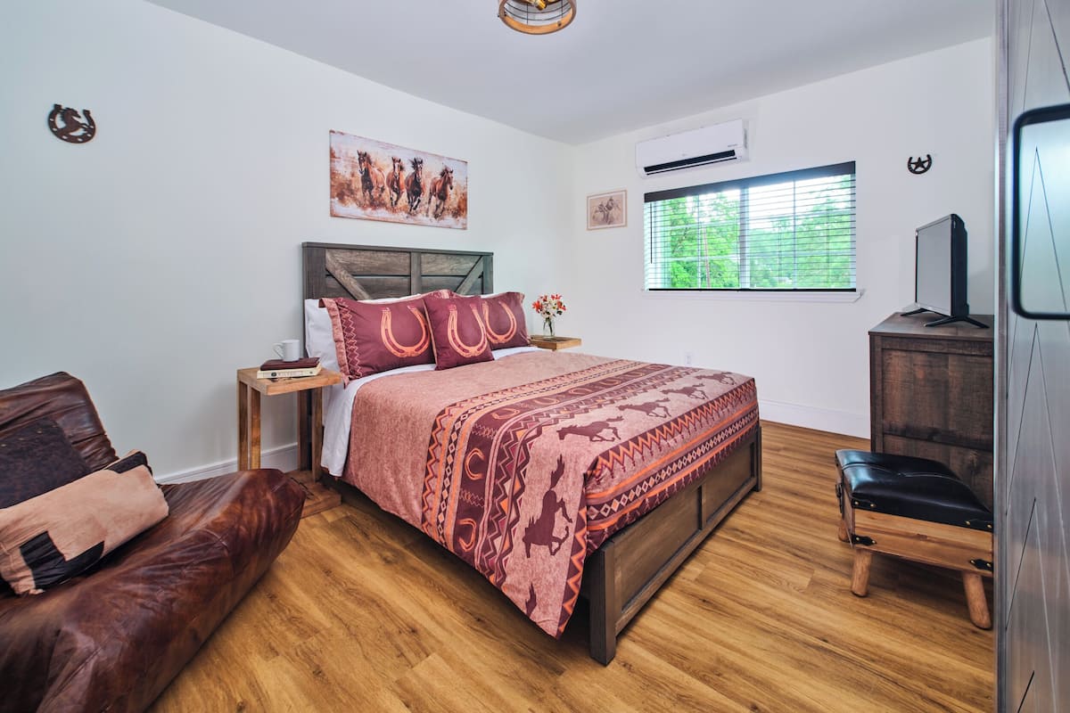 Un dormitorio con cama de madera, sofá de cuero, mesita de noche de madera, televisor y ventana con persianas.