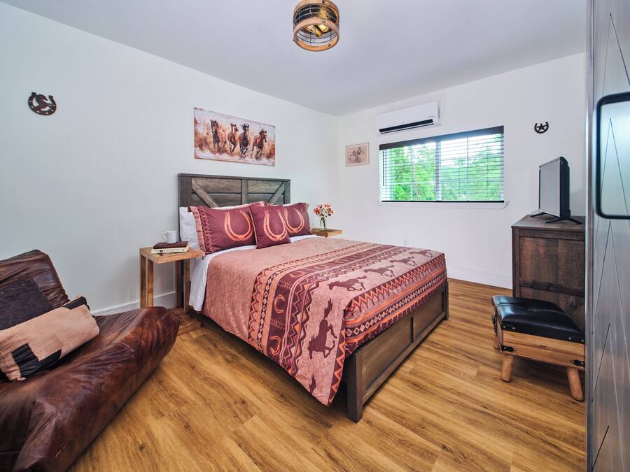 A bedroom with a wooden bed, a leather couch, a wooden nightstand, a TV, and a window with blinds.