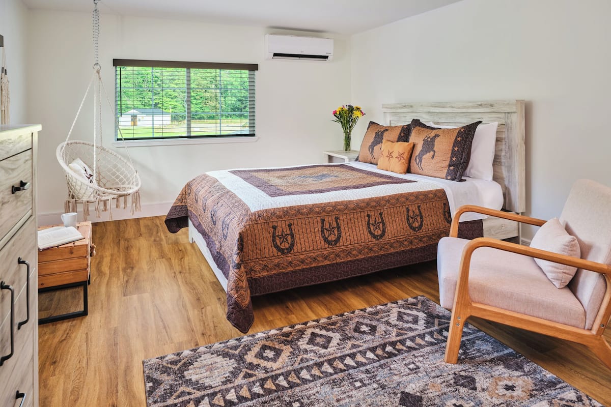 Un dormitorio con una cama grande, una silla colgante, un aparador de madera y vistas a la vegetación exterior.