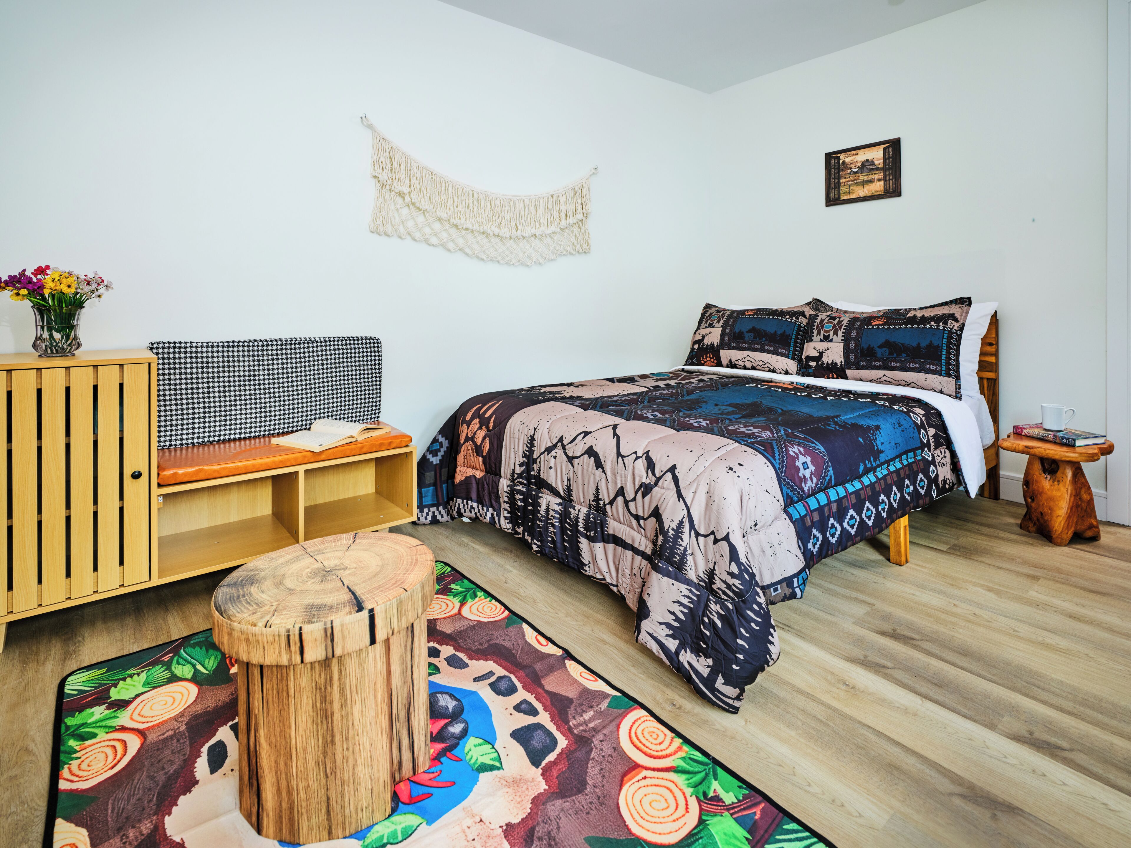A bedroom with a bed, a wooden bench, a small stool, a macrame wall hanging, and a framed picture on the wall.