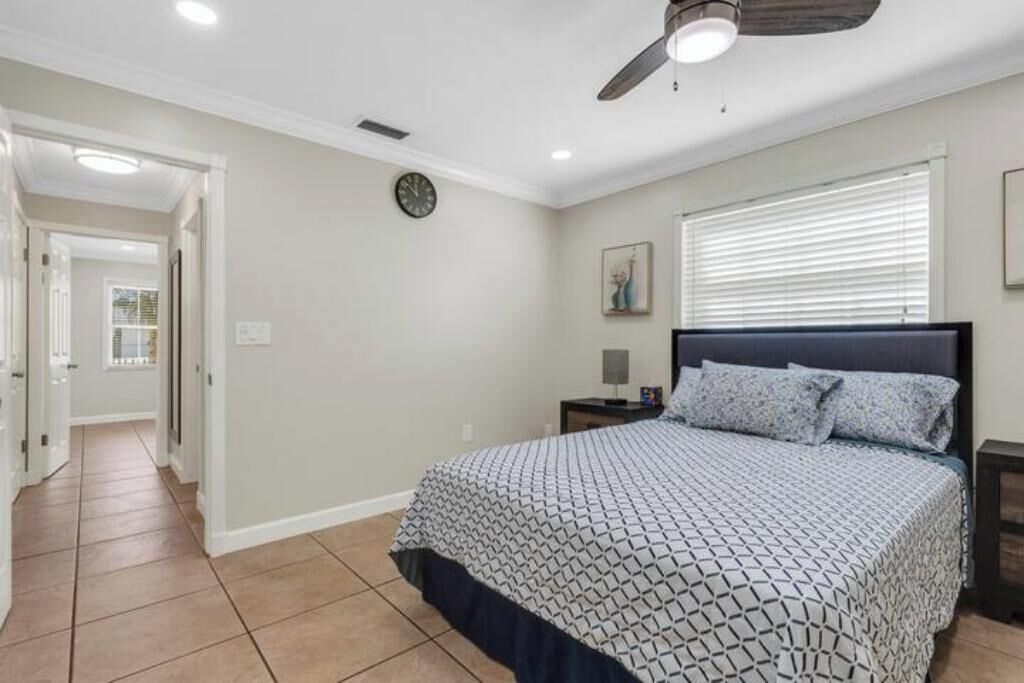Une chambre à coucher comprenant un lit, une table de chevet, un ventilateur de plafond, une horloge et une fenêtre avec des stores.