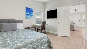 A bedroom with a bed, a desk, a chair, a TV, and a door leading to another room.