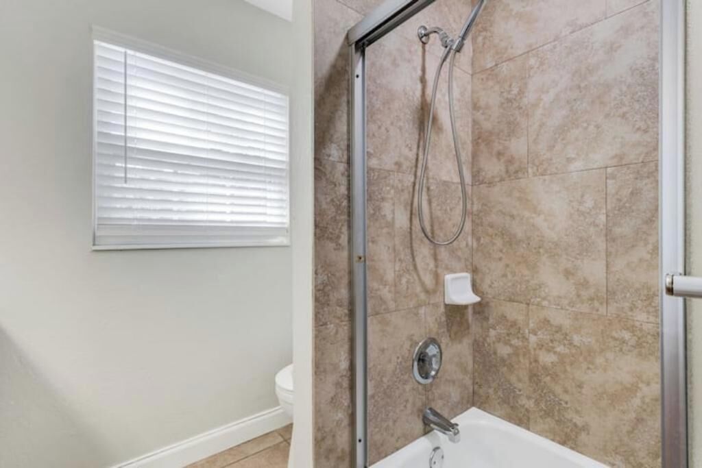 A bathroom with a shower, a toilet, and a window with blinds.