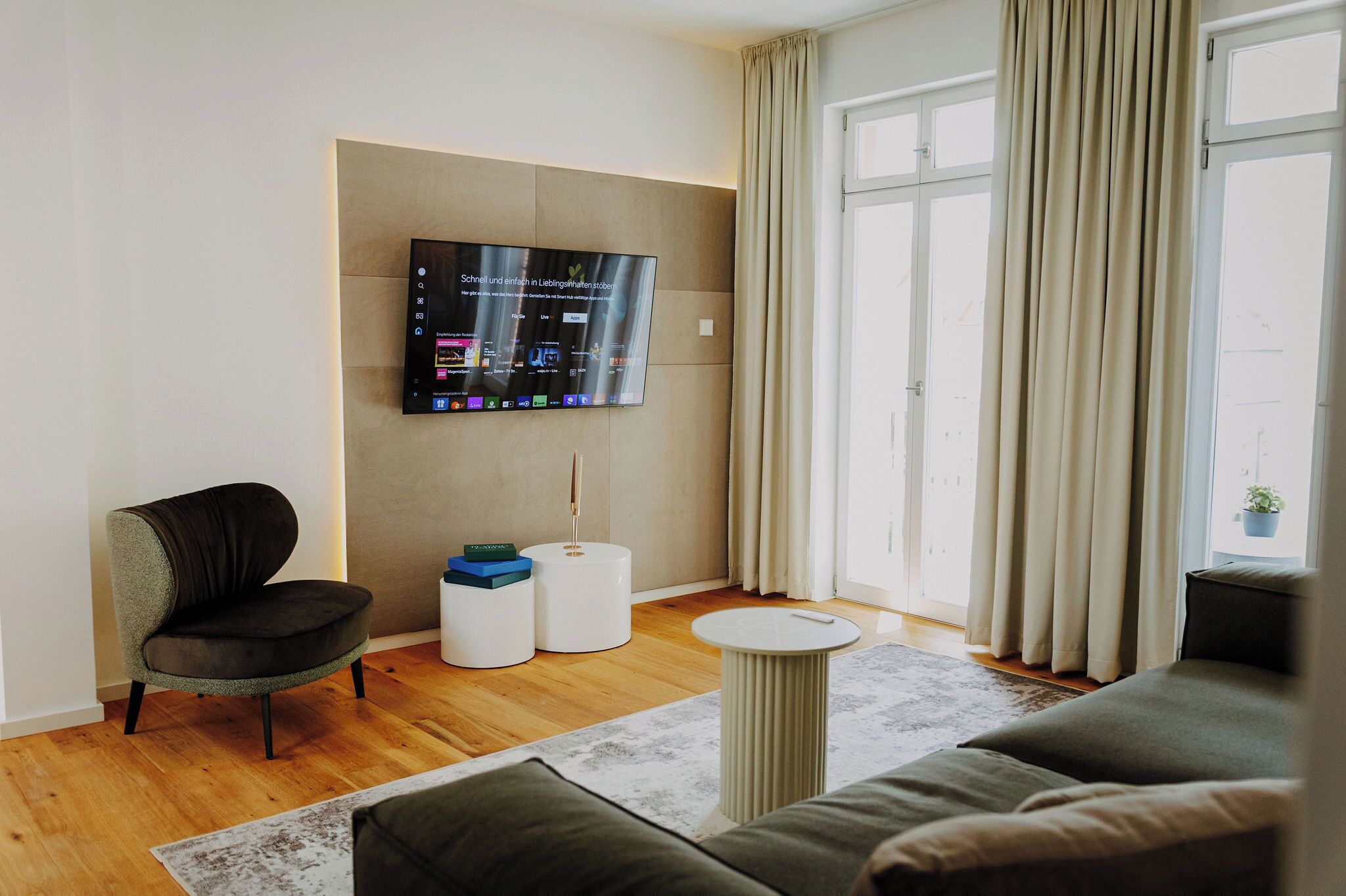 A modern living room with a flat-screen TV mounted on the wall, a comfortable sofa, a small side table, and a window with curtains.