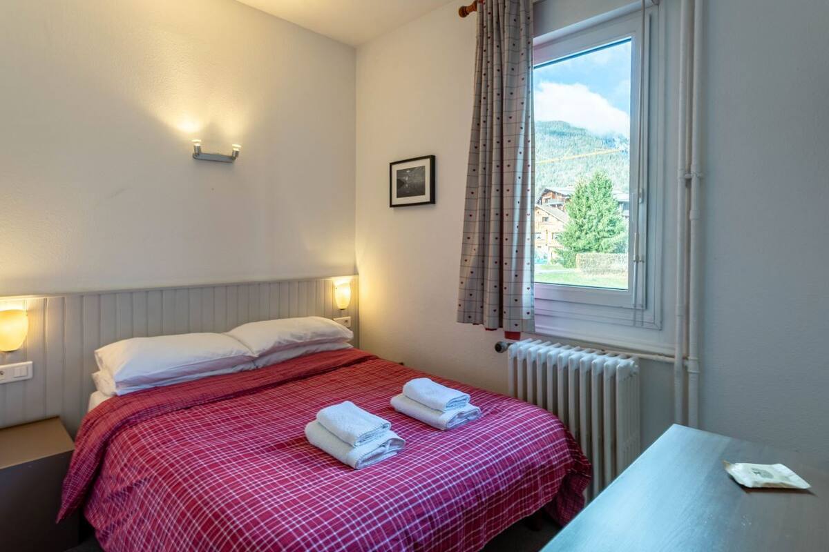 A hotel room with a bed, two bedside lamps, a window with curtains, a radiator, and a small table with a packet on it.