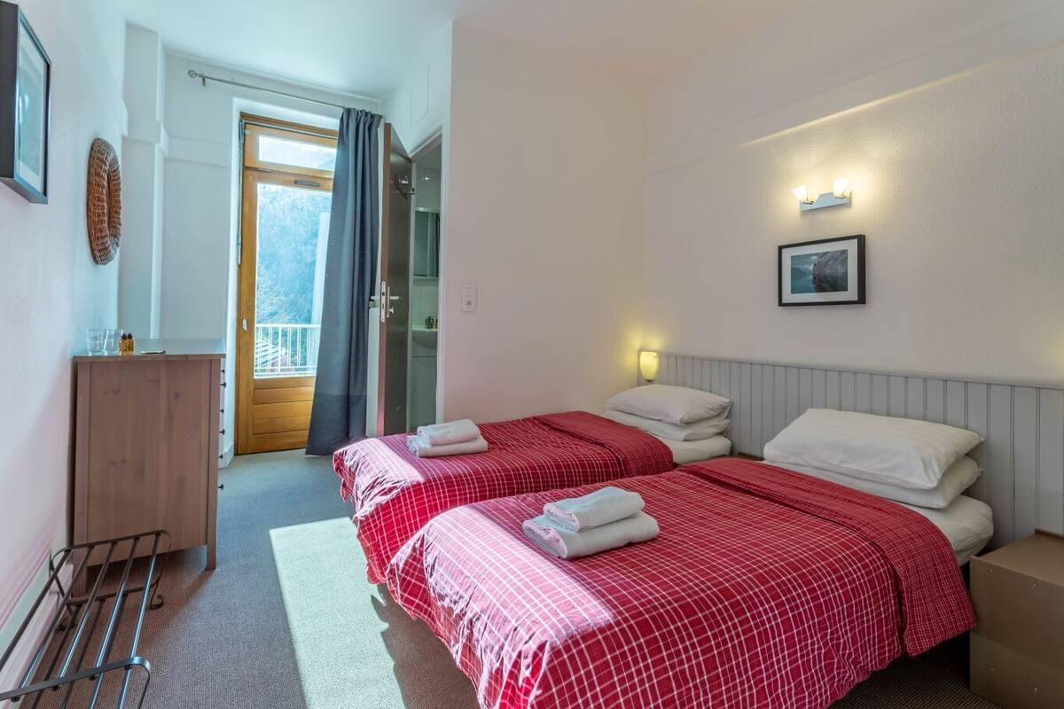 A hotel room with two beds, a wooden wardrobe, a small table, and a window with curtains.