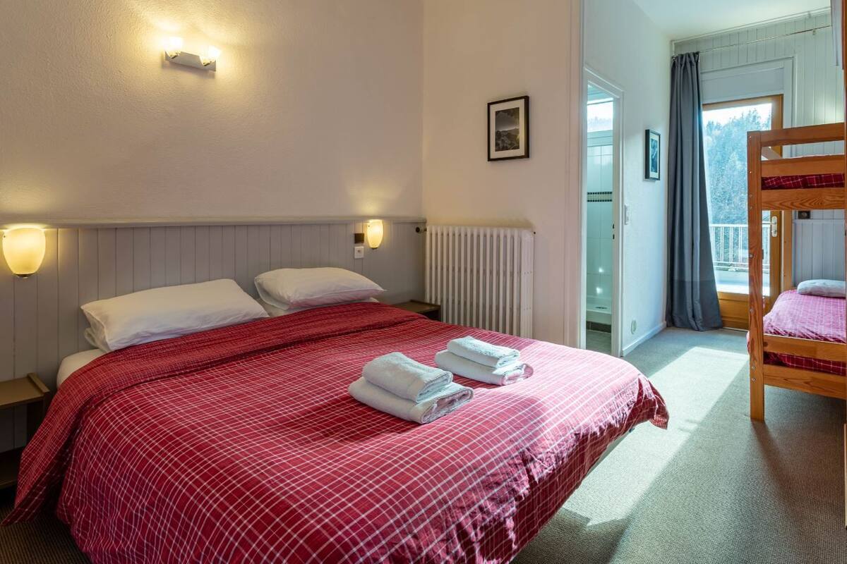 A hotel room with a large bed, two bedside lamps, a radiator, a window with curtains, and a bunk bed in the corner.