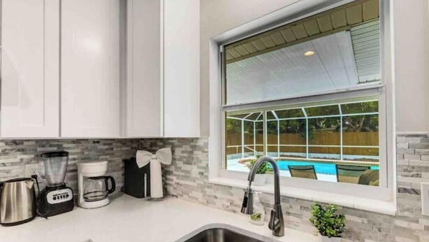A modern kitchen with white cabinets, a stainless steel sink, a coffee maker, and a window overlooking a swimming pool. - Tropical Paradise for 8 (Bradenton)
