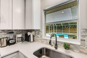 A modern kitchen with white cabinets, a stainless steel sink, a coffee maker, and a window overlooking a swimming pool. - Tropical Paradise for 8 (Bradenton)