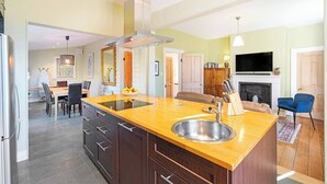 A modern kitchen with a central island, a dining area with chairs, a sofa, a fireplace, and a TV mounted on the wall. - Rose Cottage Charming Haven near CBD and Waterfront (North Hobart)