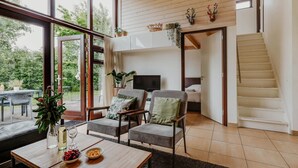 A spacious living area with a large window, a sofa, a dining table with fruit and drinks, and a staircase leading to another level. - Dutchen Vakantiepark Buiten Bergen (Schoorl)