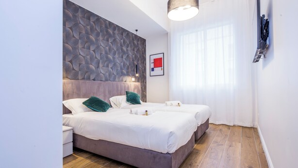 Una camera d'albergo moderna con un grande letto, una testiera effetto pietra, un comodino e una grande finestra con tende trasparenti.