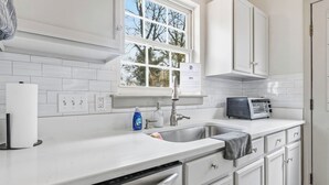 A kitchen with white cabinets, a built-in dishwasher, a sink with a faucet, a window with a view of trees, and a toaster on the counter. - Reid Cottage |3Bd/2Bth Ranch |King Beds |Sleeps 8 (Charlotte)