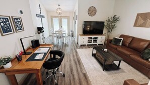 A modern living room with a wooden dining table, a brown leather sofa, a TV, and a bookshelf. - Reid Cottage |3Bd/2Bth Ranch |King Beds |Sleeps 8 (Charlotte)