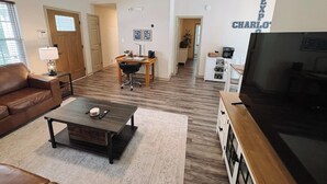 A spacious living room with a brown leather couch, a coffee table, a TV, and a dining area with a wooden table and chairs.
