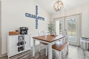 A dining area with a wooden table, white chairs, a coffee station, and a wall-mounted sign. - Reid Cottage |3Bd/2Bth Ranch |King Beds |Sleeps 8 (Charlotte)
