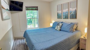 A bedroom with a bed, a TV mounted on the wall, a window with blinds, and artwork on the wall.
