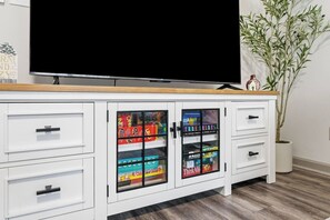 Un mueble blanco con puertas de vidrio que contiene juegos de mesa, un televisor encima y una planta en maceta a la derecha.