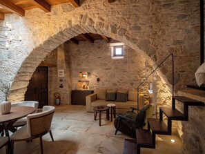 A stone-walled room with a wooden ceiling, a sofa, a chair, a small table, and a staircase. - Santa Veneranda Suites by Checkin (Minoa Pediada)
