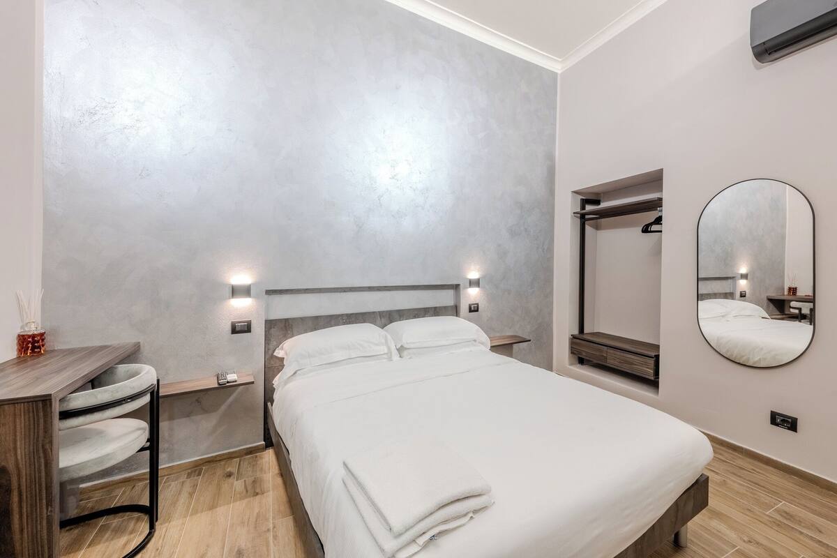 A modern hotel room with a bed, bedside tables, a mirror, and a small seating area.