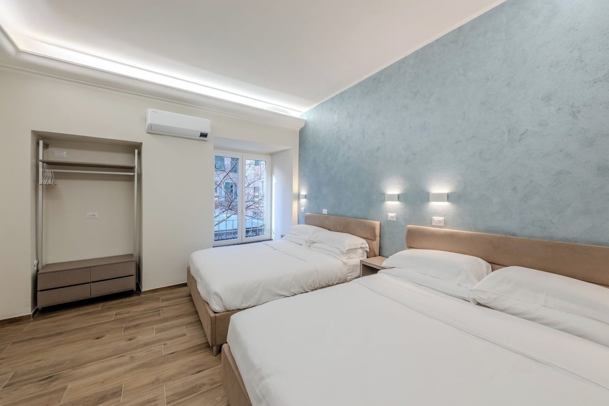 A hotel room with two beds, a wardrobe, an air conditioning unit, and a window with a view of buildings.