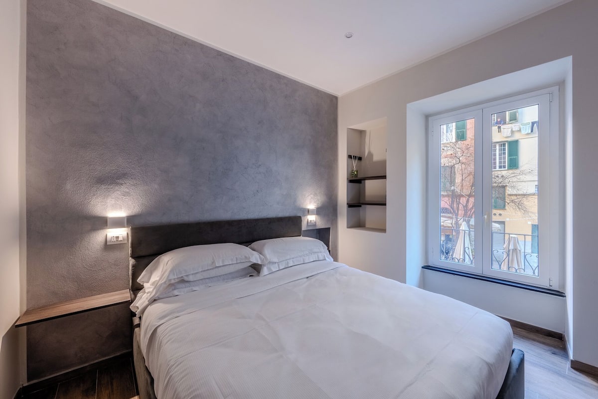 A modern bedroom with a large bed, a grey textured wall, and a window with a view of buildings.