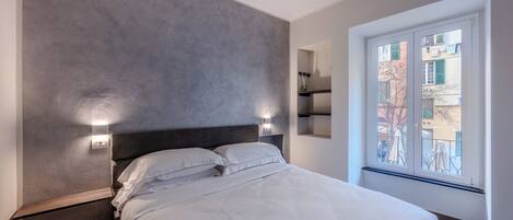 A modern bedroom with a large bed, a grey textured wall, and a window with a view of buildings.