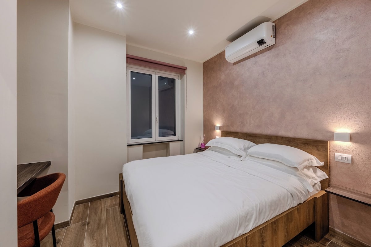 A bedroom with a wooden bed, a window with blinds, a desk with a chair, and a wall-mounted air conditioner.