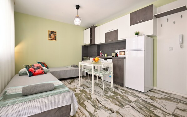 Habitación compacta con cama, una pequeña mesa de comedor con sillas, kitchenette con refrigerador y microondas.