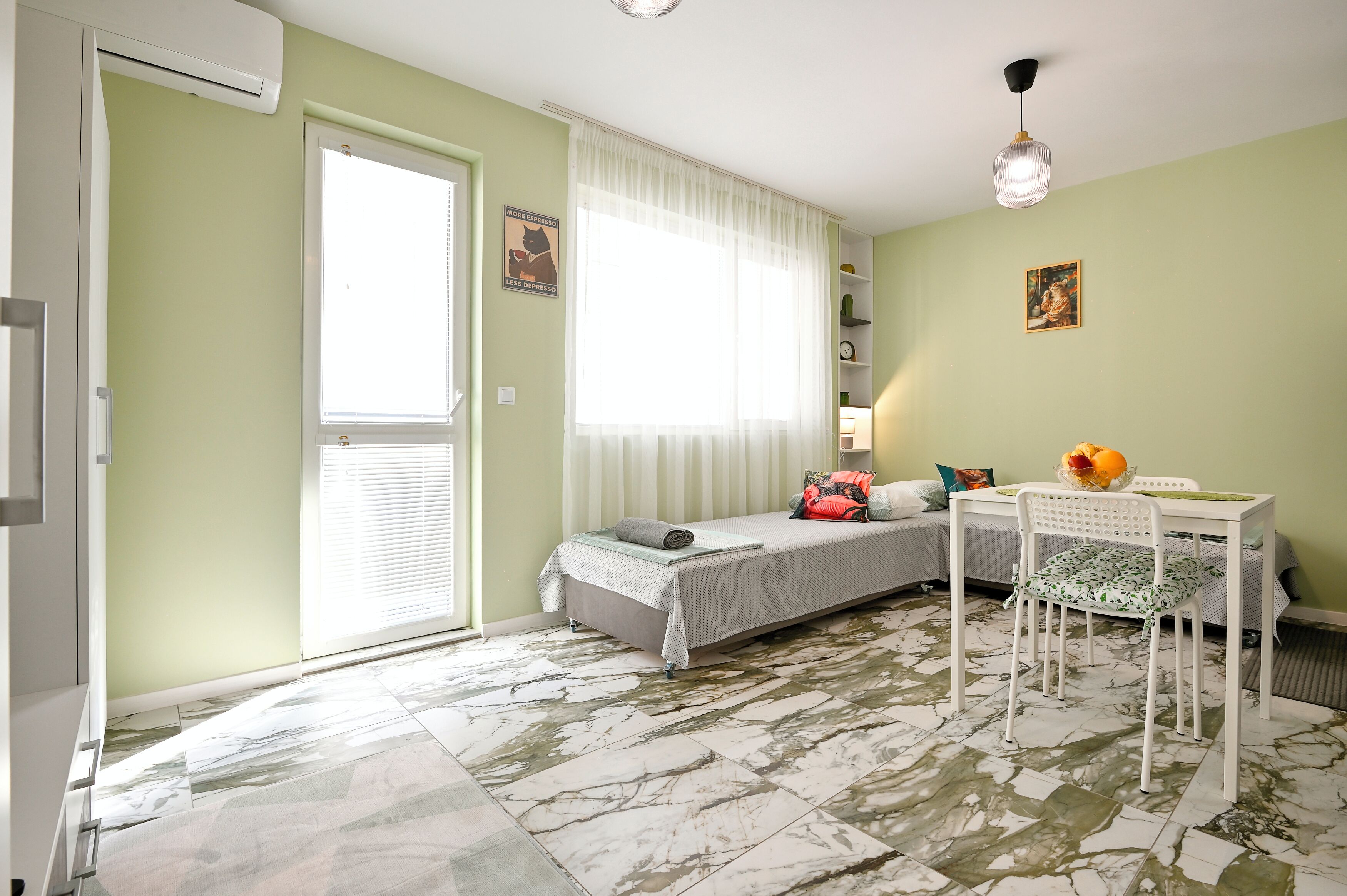 A room with a bed, a table with a chair, a radiator, a window with curtains, and a picture on the wall.