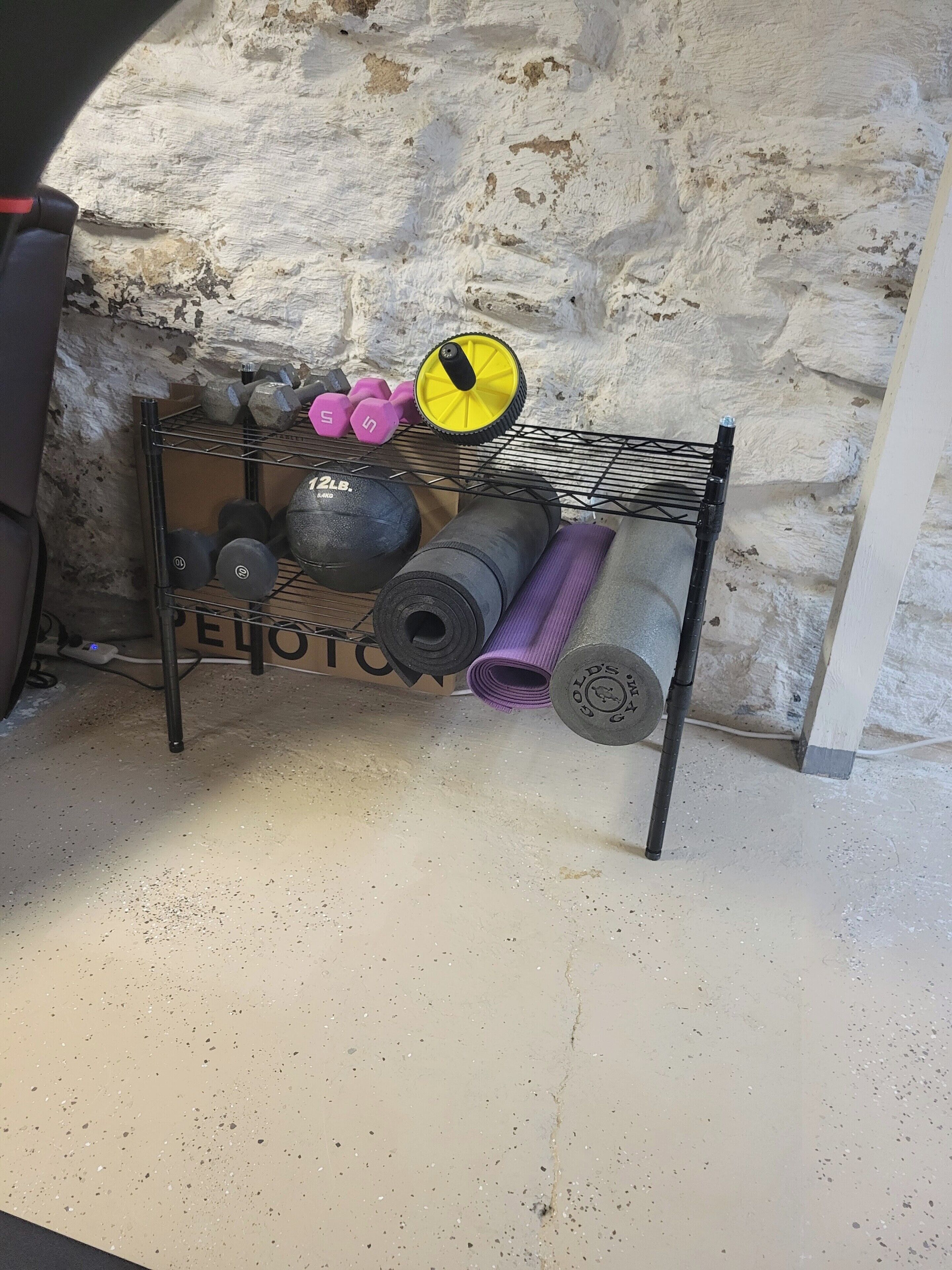 Una prestatgeria metàl·lica amb peses, un matalàs de ioga i una kettlebell davant d'una paret amb relleu.
