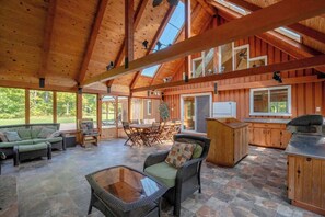 A spacious indoor area with high ceilings and wooden beams, featuring a dining area with chairs and a table, a sofa, and a kitchen area with a counter and cabinets. - Outdoor Kitchen, BBQ, Fire Pit & Hot Tub (Saint-Raymond)
