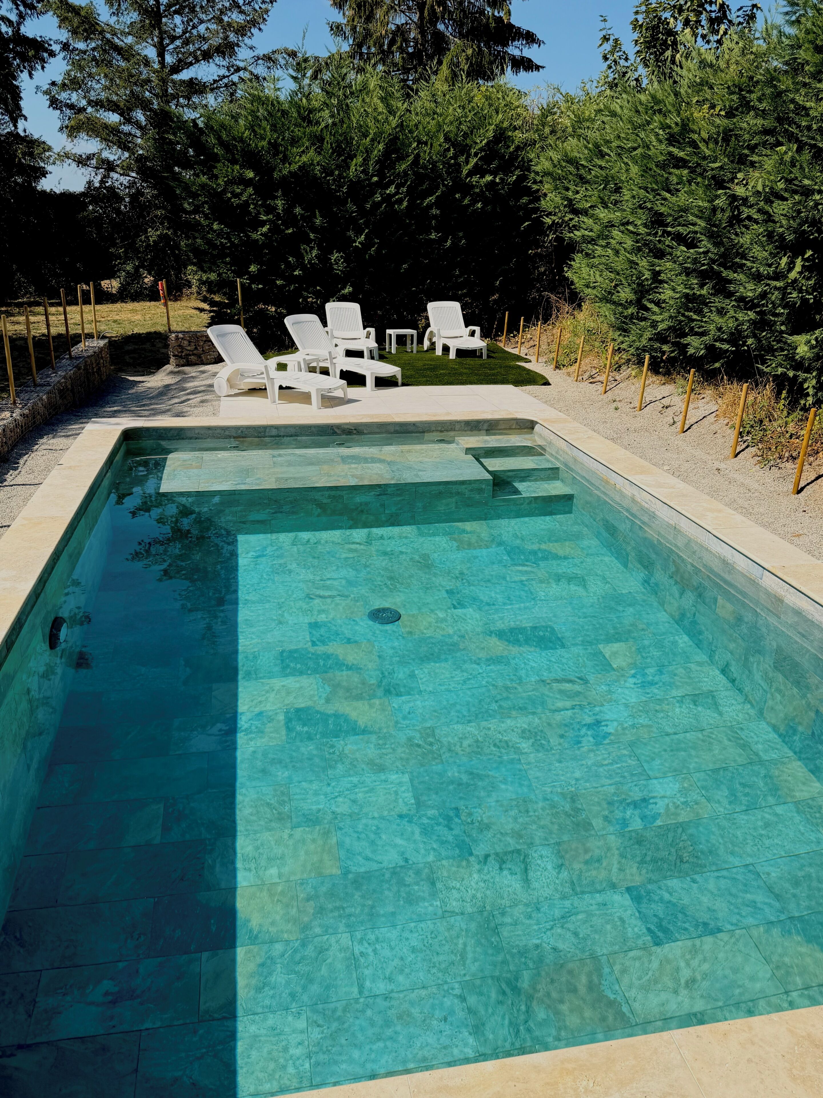 A rectangular swimming pool with clear water, surrounded by a stone deck. There are white lounge chairs and a small table near the pool's edge.