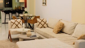 Een moderne woonkamer met een beige bank, een ronde salontafel, en een eethoek met een kleine tafel en stoelen.