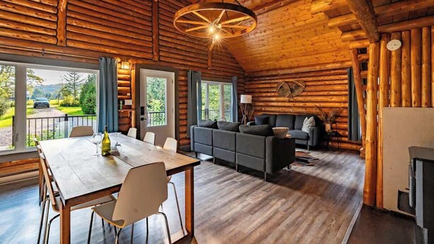 A log cabin interior with a dining area, sofa, and large windows offering a view of the outdoors.
