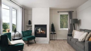 Een moderne woonkamer met een open haard, groene en grijze banken, een klein bijzettafeltje en een vloerlamp.