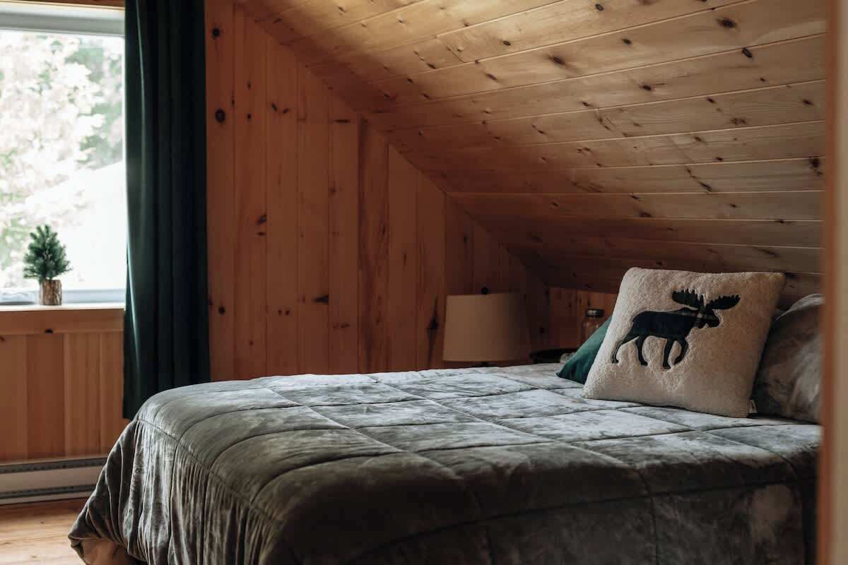 Een knus ingerichte kamer met een houten plafond, een bed met een gebogen deken, een kussen in de vorm van een eland en een kleine plant bij het raam.