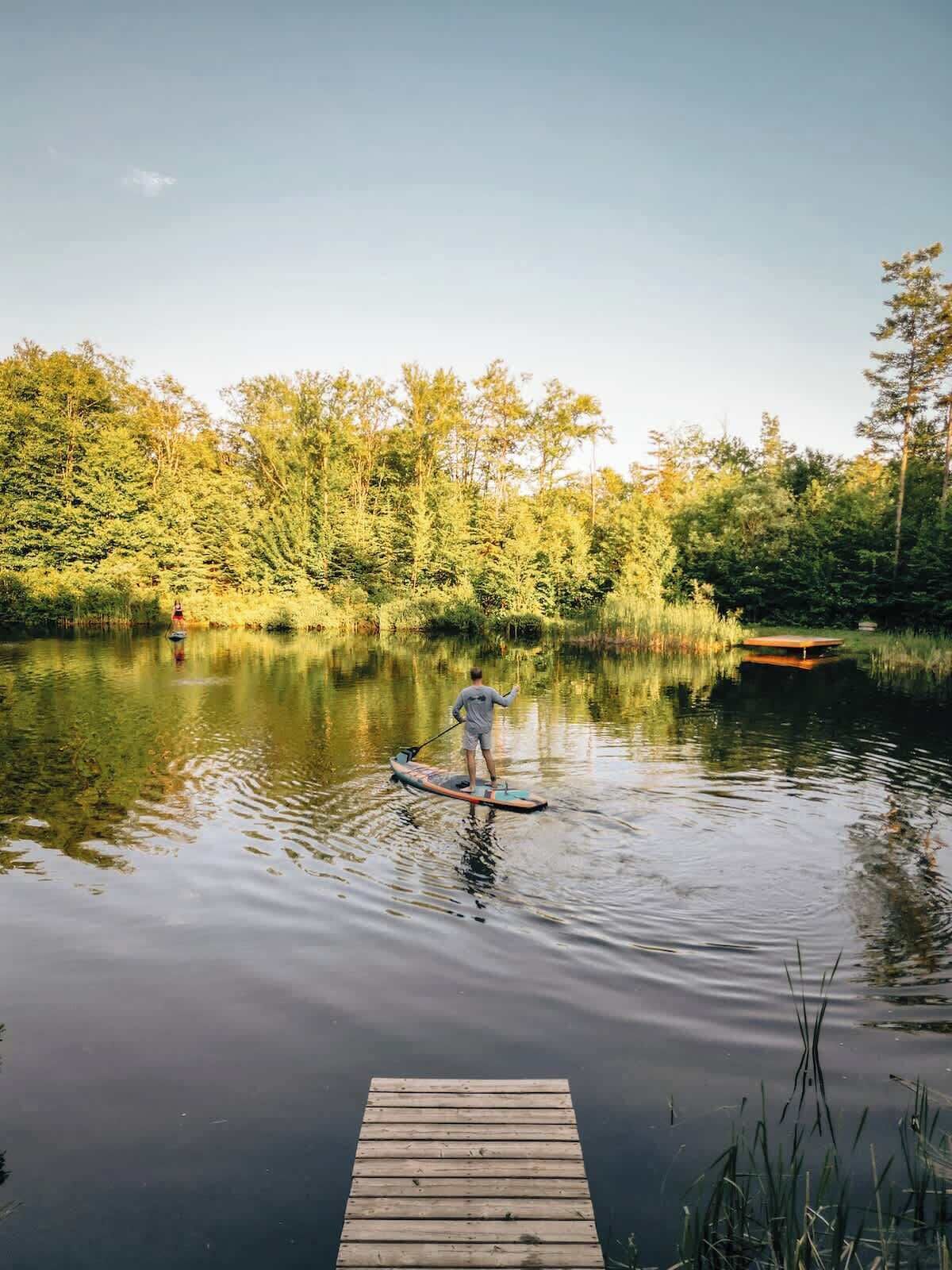 Eine Person steht auf einem Stand-Up-Paddle auf einem ruhigen See, im Vordergrund ein Holzsteg und im Hintergrund dichtes Baumbestand.