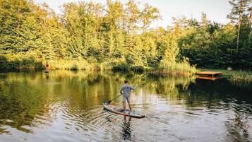 Eine Person steht auf einem Stand-Up-Paddle auf einem ruhigen See, im Vordergrund ein Holzsteg und im Hintergrund dichtes Baumbestand.