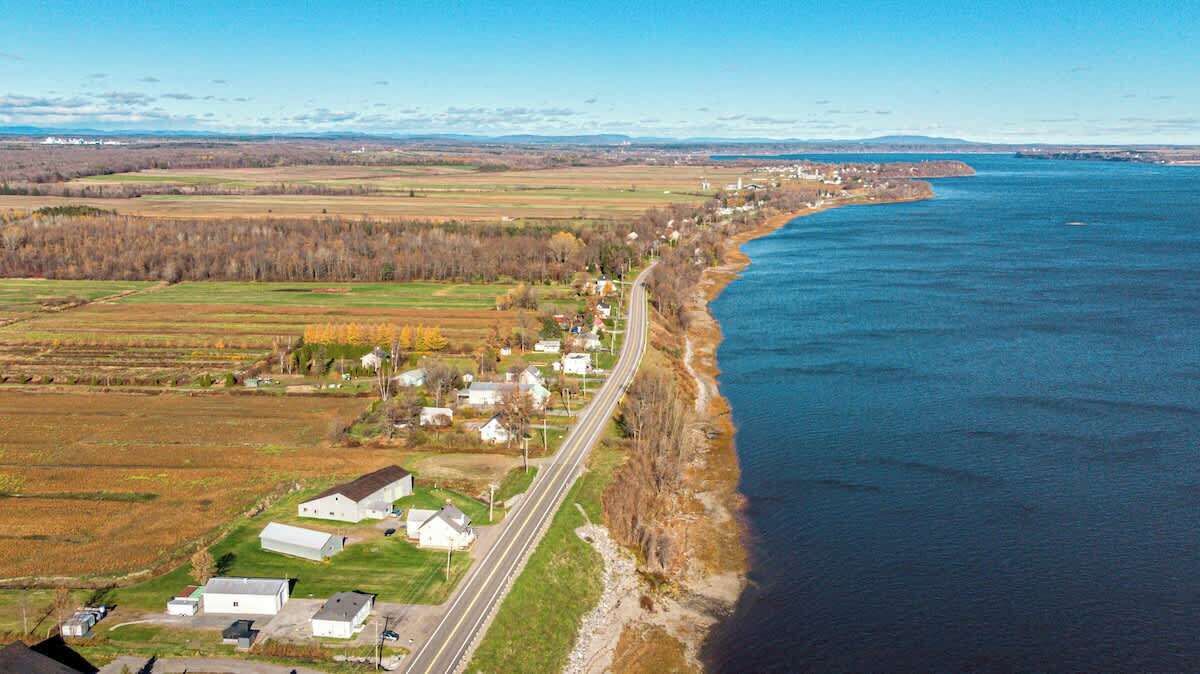 An aerial view of a coastal town with a road running parallel to the shoreline, houses, and open fields.
