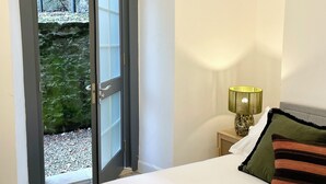 Un dormitorio con cama, una mesita de noche con lámpara, ventana con vistas a un muro de piedra y follaje, y una puerta que da al exterior.