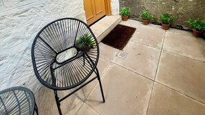 Un edificio con pared blanca texturizada, una puerta de madera con panel de vidrio y el número 59A. Hay tres macetas con plantas en el suelo y una silla de metal negro al frente.