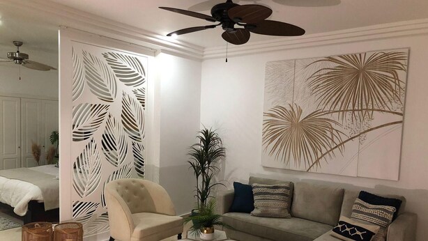 Una sala de estar con sofá, sillón y mesa de centro. Hay un ventilador de techo, un panel decorativo en la pared y una planta grande.