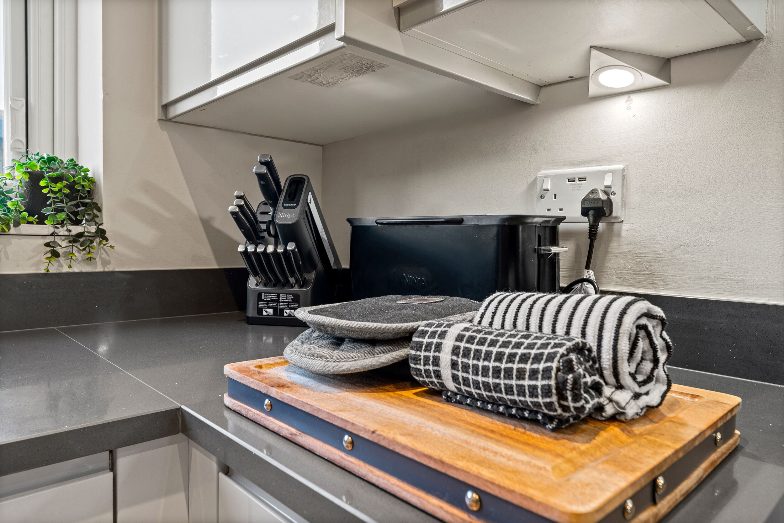 A kitchen counter with a wooden cutting board, a black kettle, and a black coffee maker.