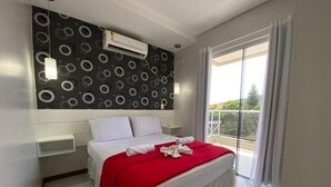 A hotel room with a bed, a red blanket, a white pillow, a wall-mounted air conditioner, and a balcony with a view. - Beautiful 1 bedroom apartment with balcony and barbecue 70m from Praia dos Ingleses (Ingleses Sul Florianópolis)