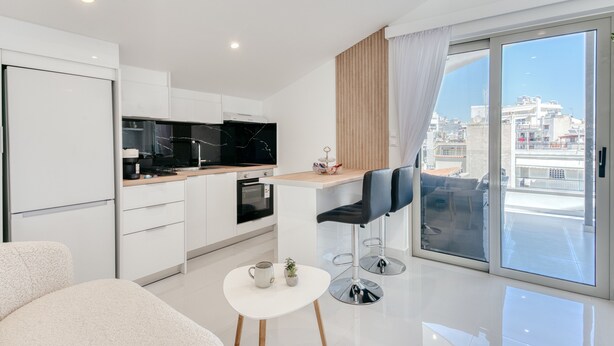 A modern kitchen with white cabinets, a black marble countertop, and a dining area with a white table and two black chairs. - Ayla Studio Athens (Imittos)