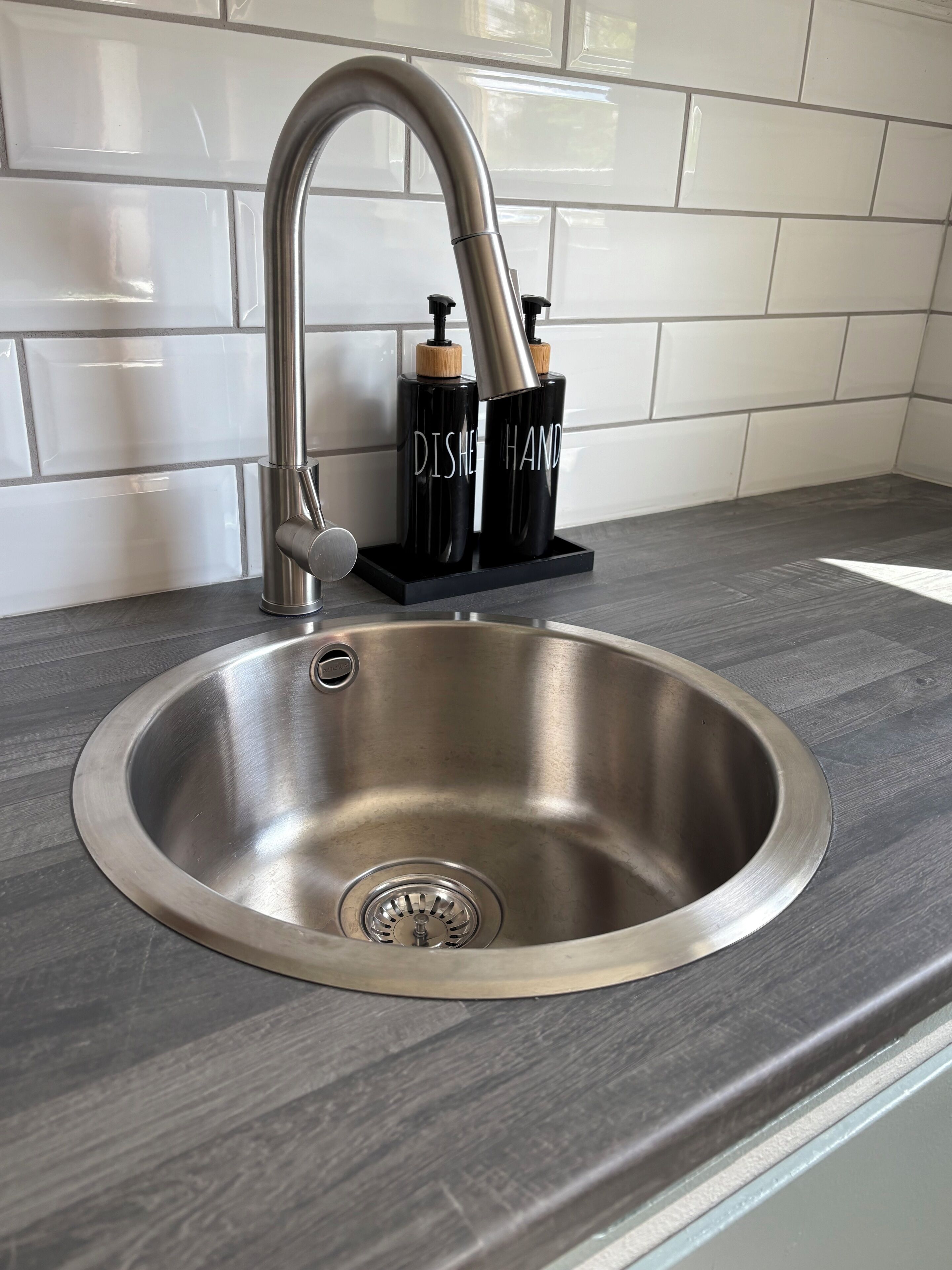 Ein Edelstahl-Spülbecken mit modernem Wasserhahn, zwei schwarzen Seifenspendern und einer grauen Arbeitsplatte.