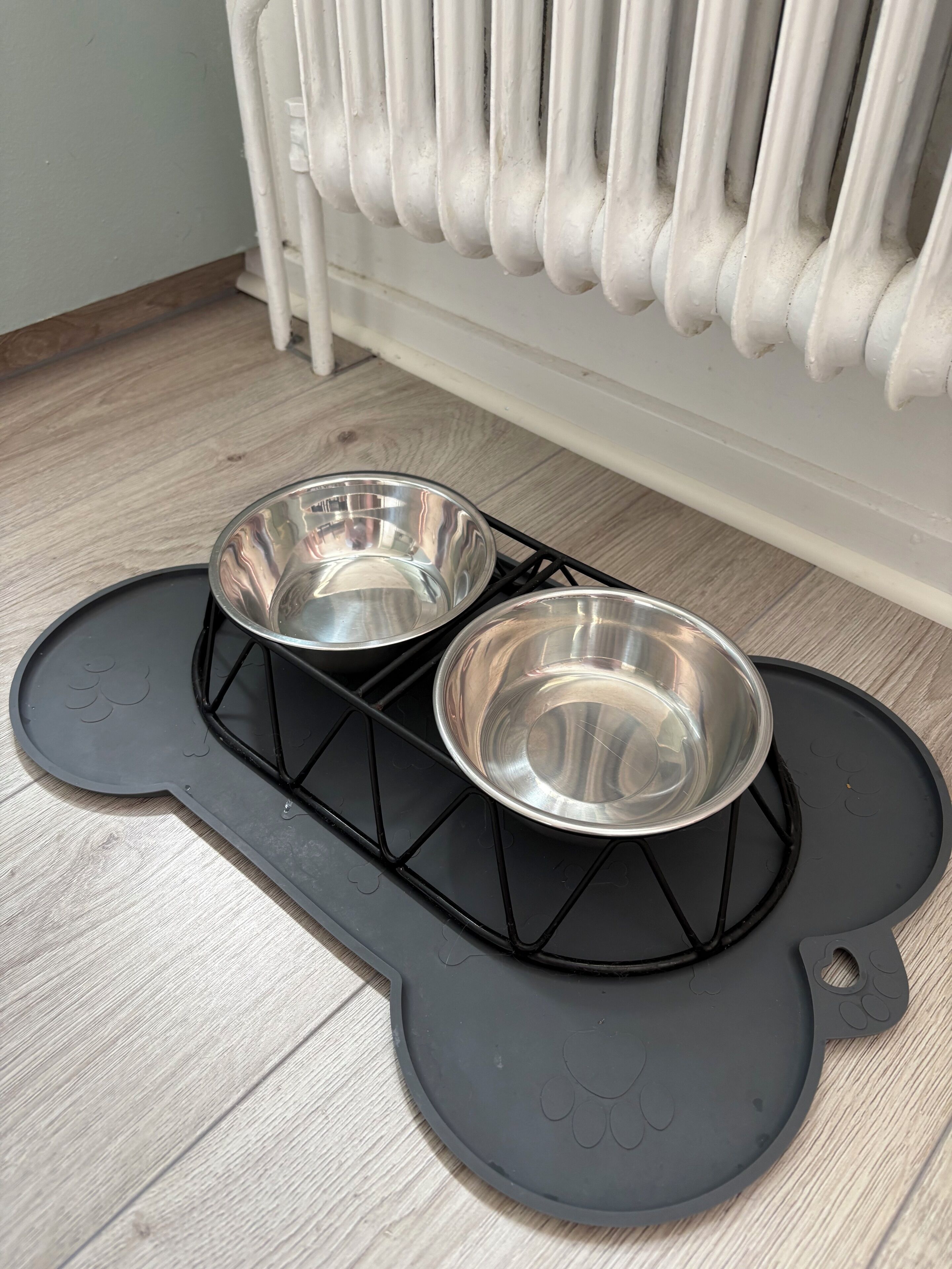A pet feeding station with two stainless steel bowls on a black mat with paw prints, placed on a wooden floor near a white radiator.